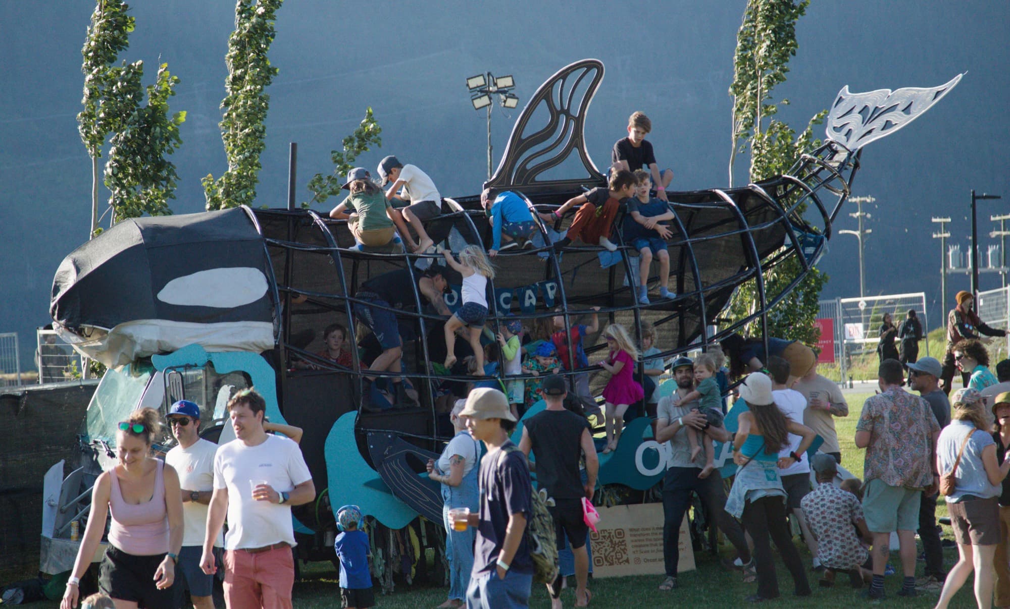 The Orcar in daylight surrounded by a festival crowd, kids climbing its steel ribs
