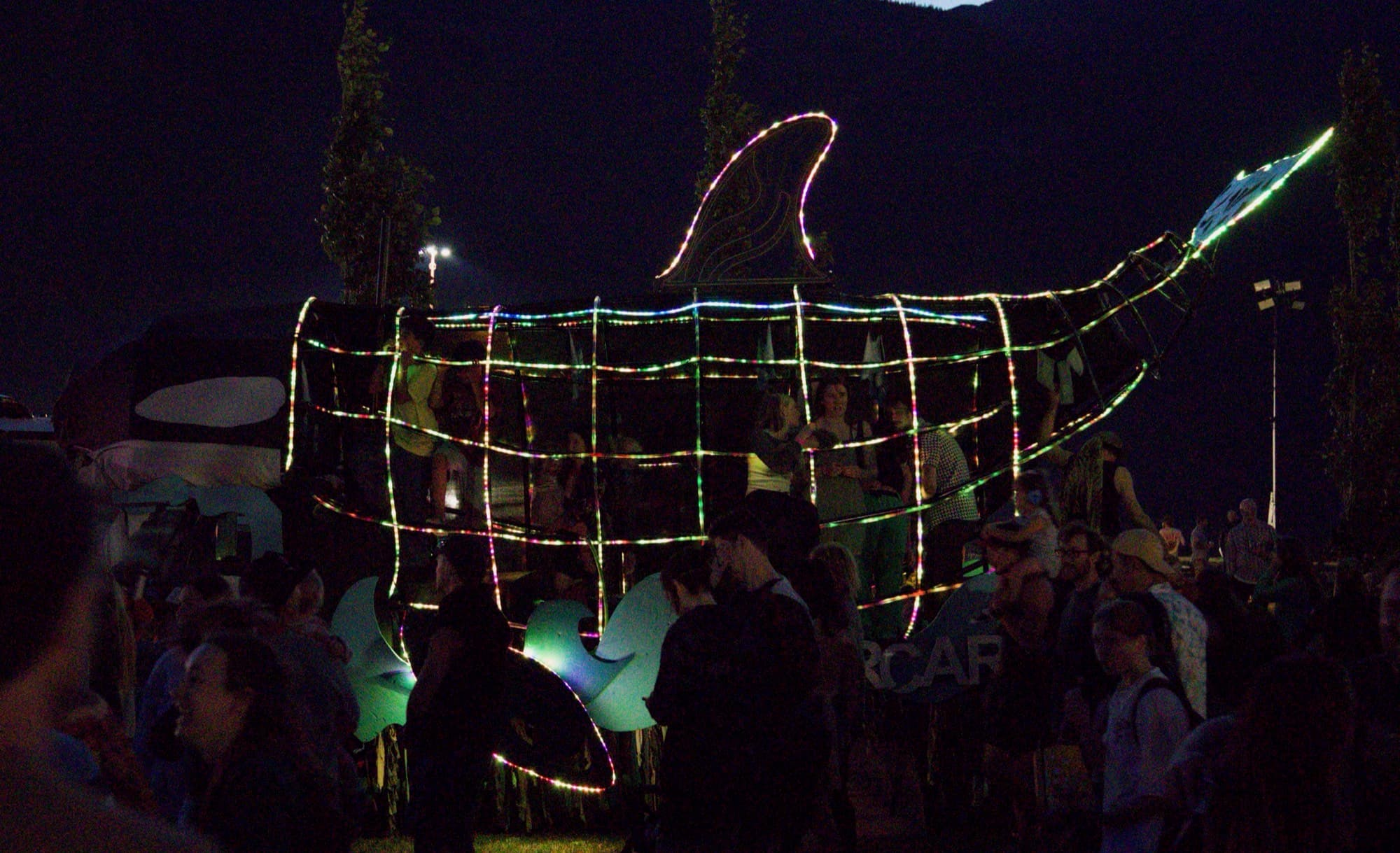 The Orcar at night with its frame traced in green and red LED rope, people standing inside the body